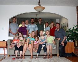 Family Fun/ Photo Op A group of 17 people, including adults and children, gather in a living room. Some are seated on a sofa, while others stand behind it. The room has a brick wall, a plant, and a wooden side table.