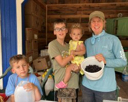 A woman and three children stand inside a wooden shelter. She holds a bucket of blackberries. The older boy holds the youngest child, while the other boy stands near a white bottle.